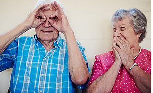 Ein Senior formt mit seinen Händen eine Brille, durch welche er schaut. Eine daneben sitzende Seniorin schaut lächelnd zu ihm herüber.